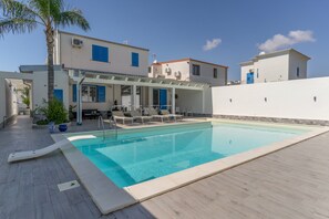 A swimming pool with lounge chairs and a pergola, surrounded by a white building with blue shutters.