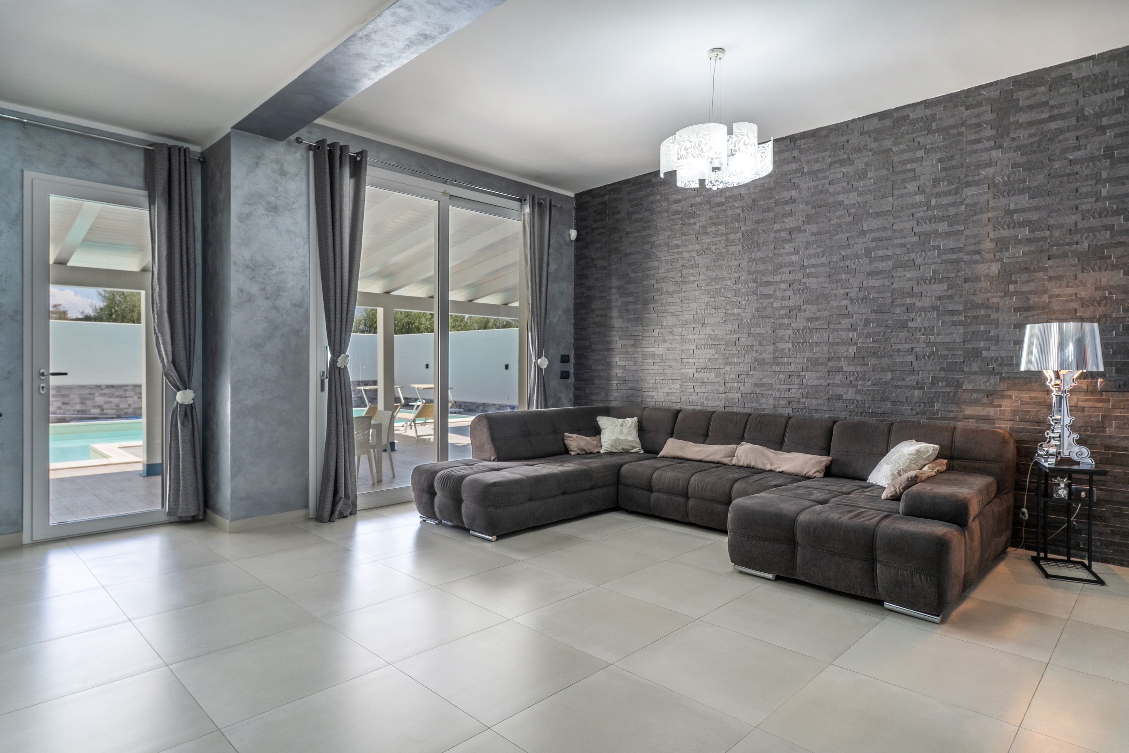 A modern living room with a sectional sofa, a chandelier, and a view of a pool through large sliding doors.