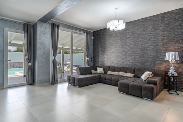 A modern living room with a sectional sofa, a chandelier, and a view of a pool through large sliding doors.