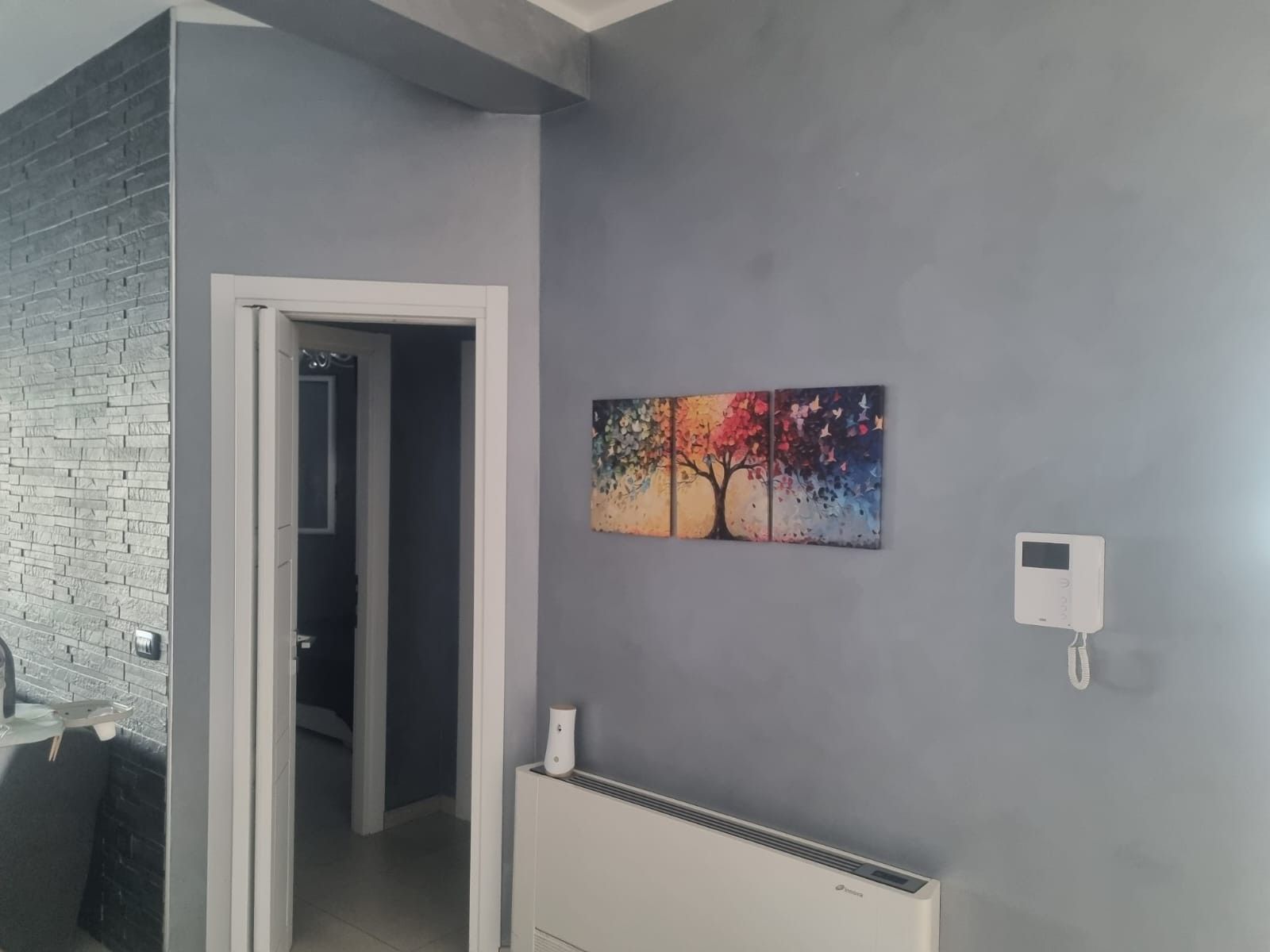 Una habitación con pared gris, una puerta que da a otra estancia, un radiador y un termostato fijado a la pared.