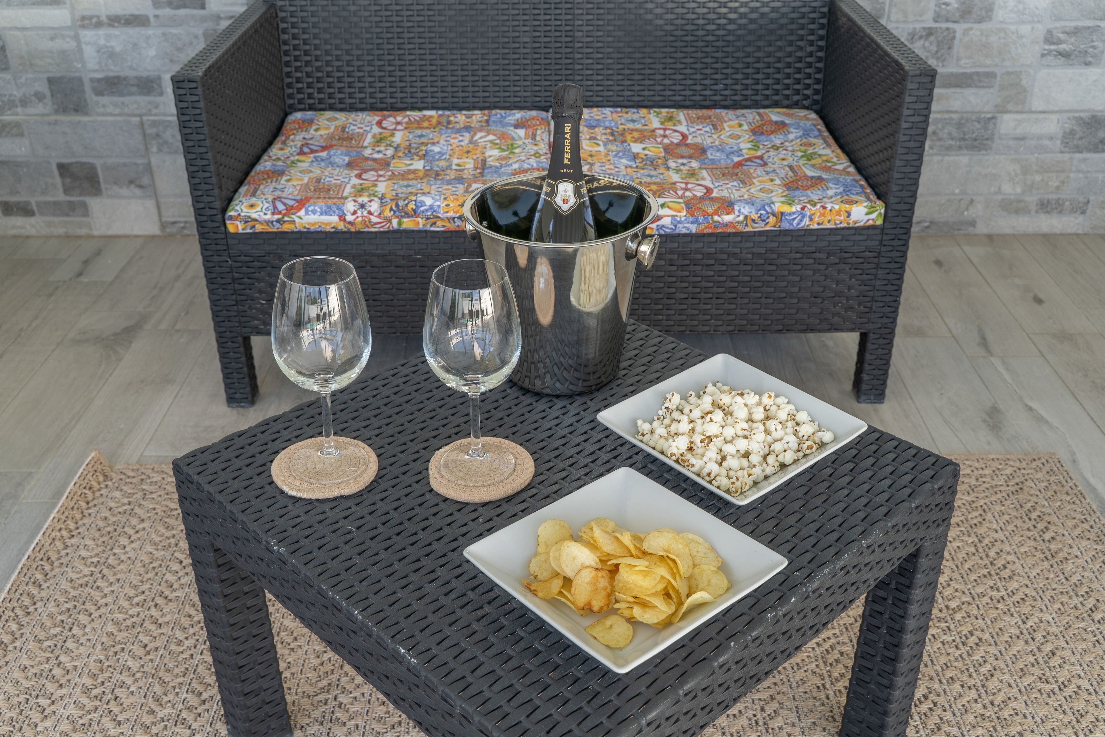 A black wicker table with two wine glasses, a bottle of champagne, and two bowls of snacks.