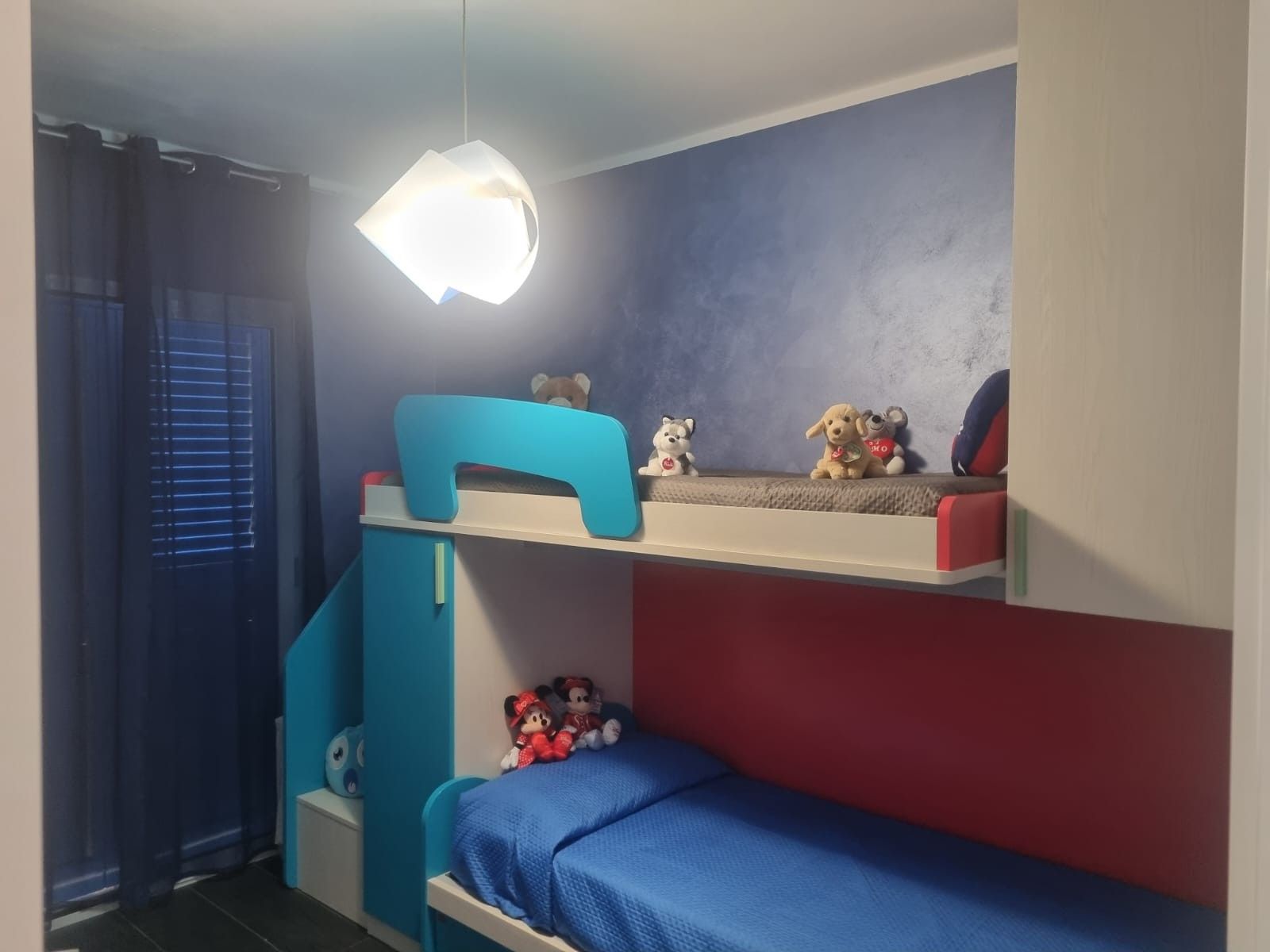 Ein Etagenbett mit Schreibtisch und Regalen, eine Hängeleuchte und ein Fenster mit Jalousien.