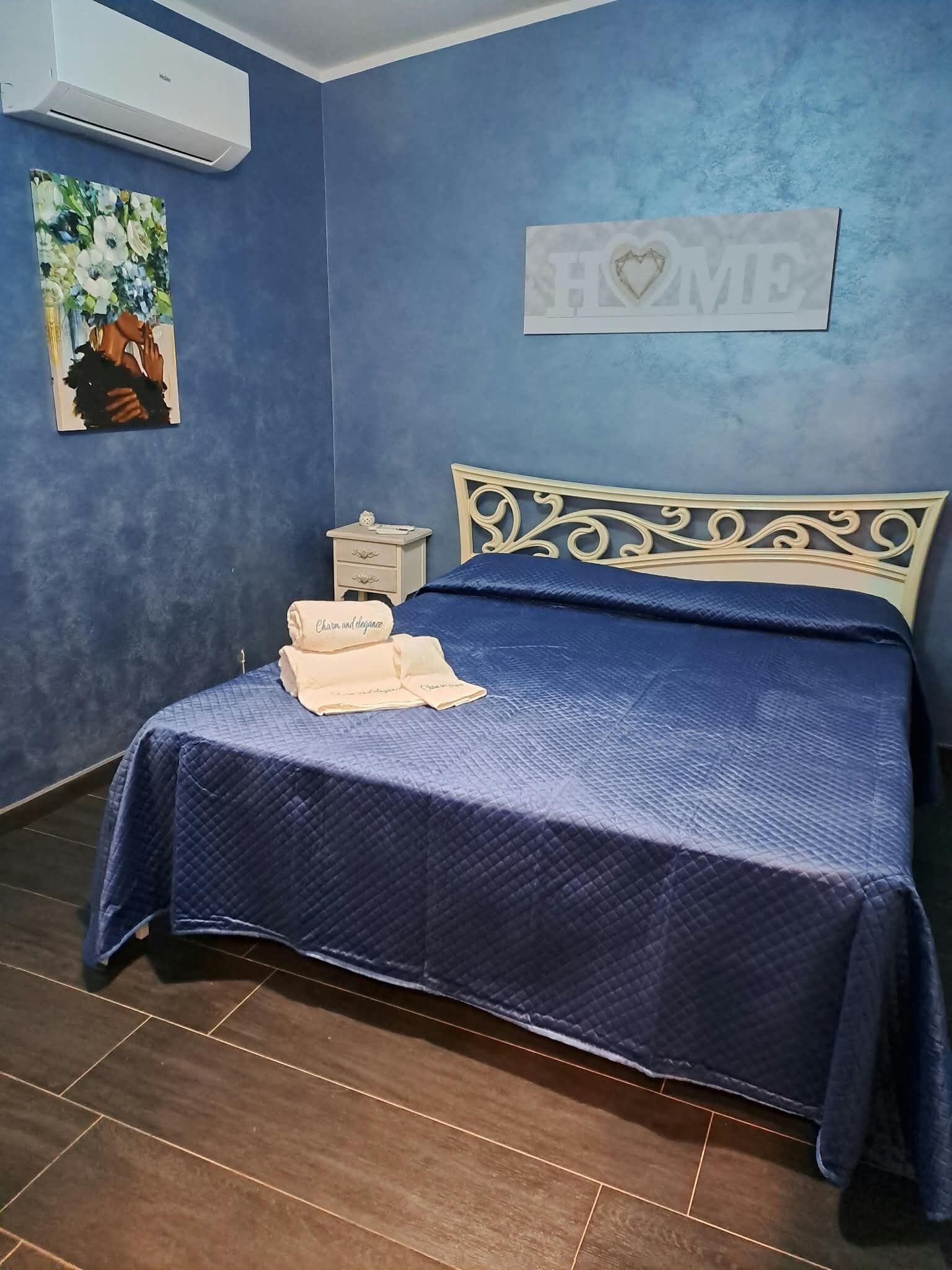 Ein Schlafzimmer mit blauer Wand, einem Bett mit dekorativer Kopfteil, einem Nachttisch mit Lampe und einem Gemälde.