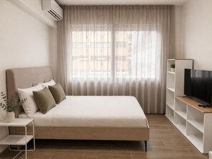Quarto moderno com cama, TV sobre suporte e janela com cortinas.