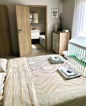 Ein Schlafzimmer mit einem Bett, einem Nachttisch mit Handtüchern, einem Babybett, einem Kleiderschrank und einem durchgehenden Türrahmen einsehbaren Badezimmer.
