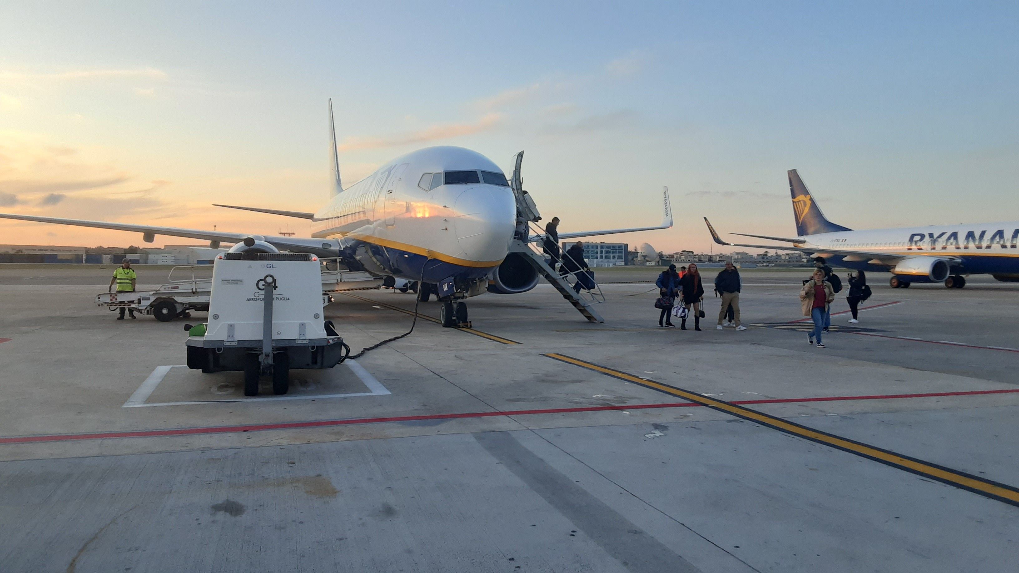 An airport tarmac with a parked airplane, ground service vehicles, and passengers boarding.