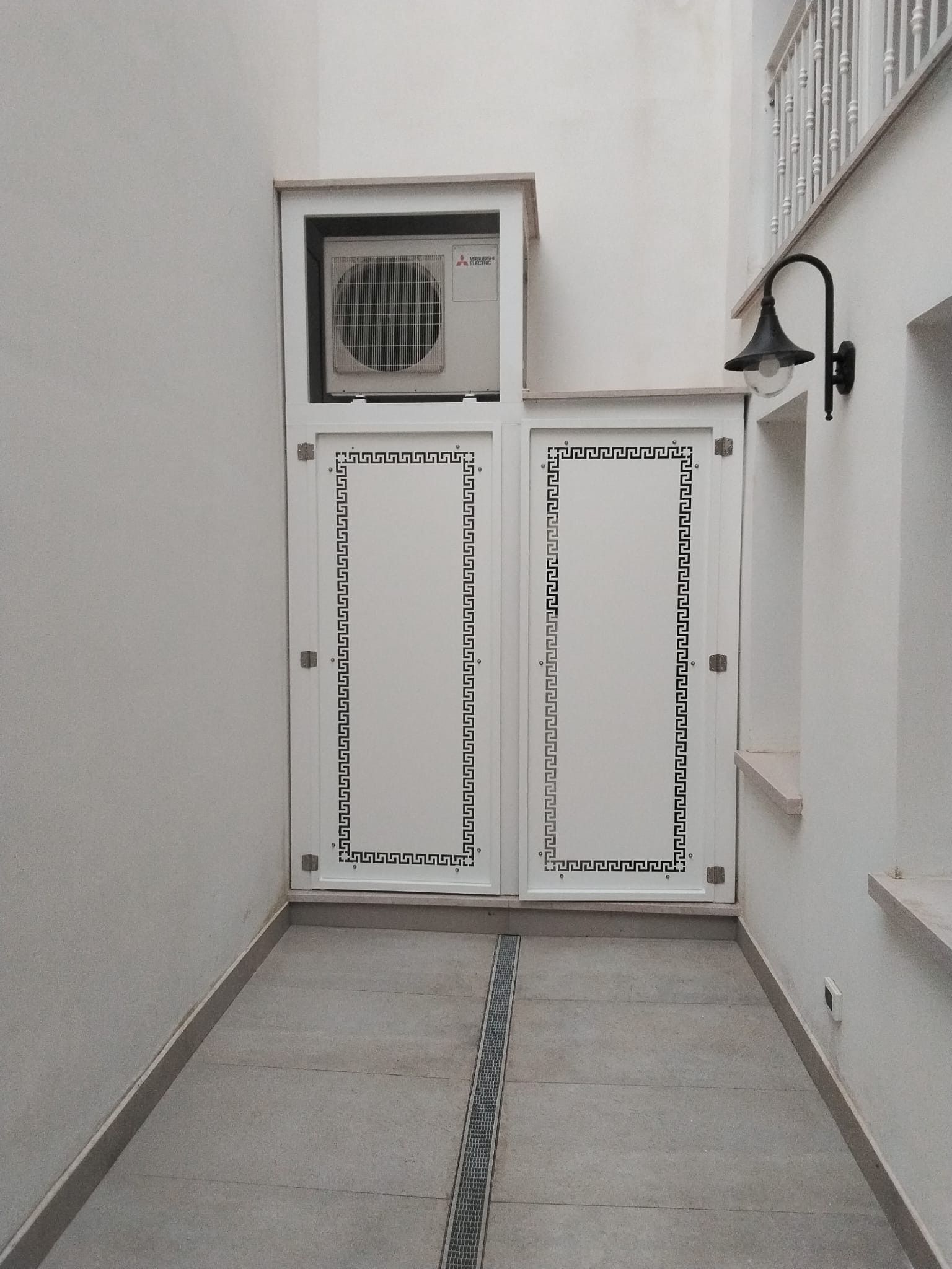 Ein Flur mit einer Doppeltür, die ein geometrisches Muster zeigt, einer Lüftungseinheit darüber und einer Wandleuchte.