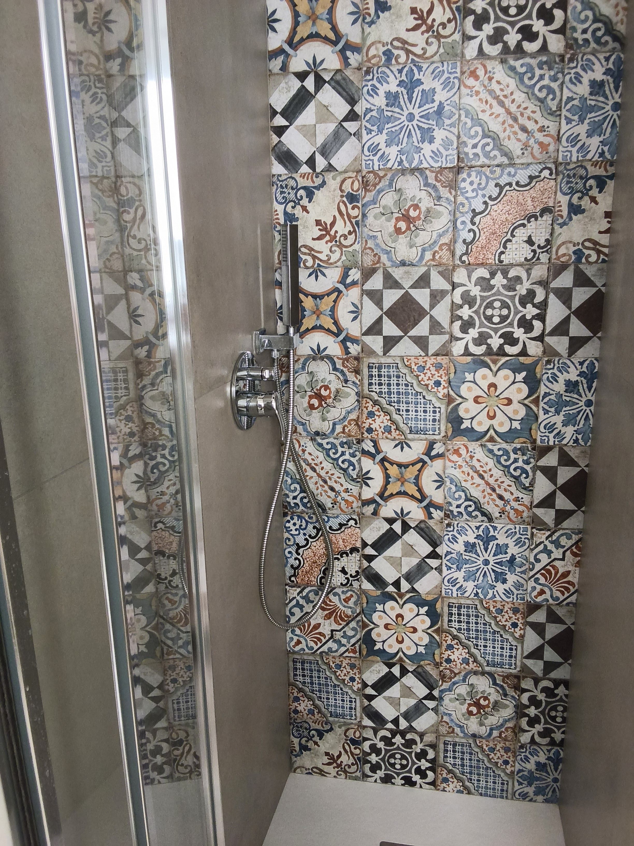 Un angolo doccia con porta in vetro e parete piastrellata decorata con motivi geometrici e floreali.