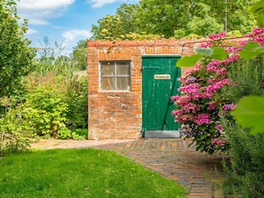 A small brick building with a green door and a window, surrounded by lush greenery and flowering plants. - Holiday Home in Upleward With Sauna (Krummhoern)