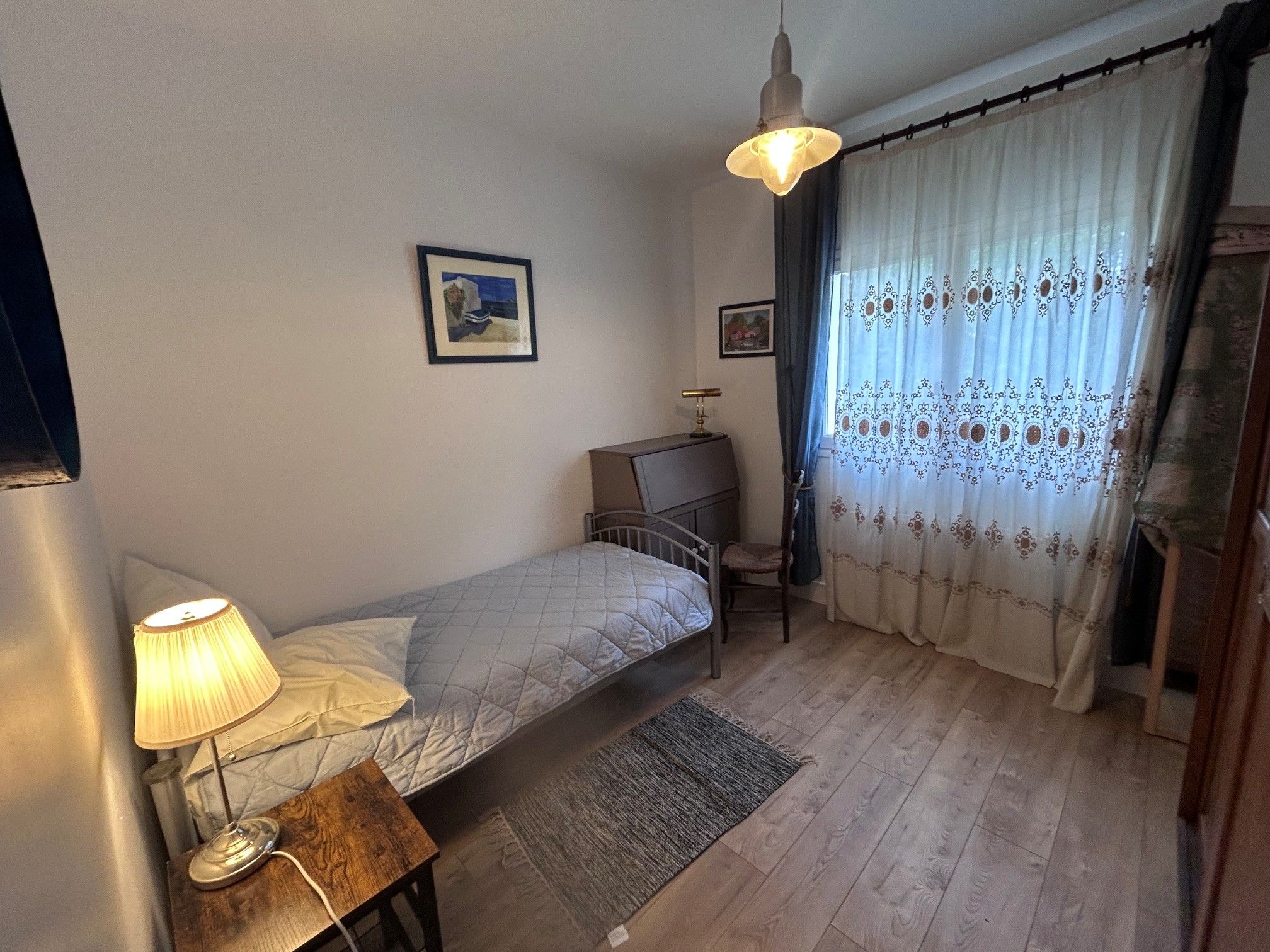 Een slaapkamer met een enkel bed, een bureau, een stoel, een lamp en een raam met gordijnen.