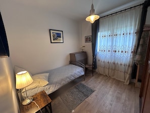 A small bedroom with a single bed, a desk, a chair, a lamp, and a window with curtains.