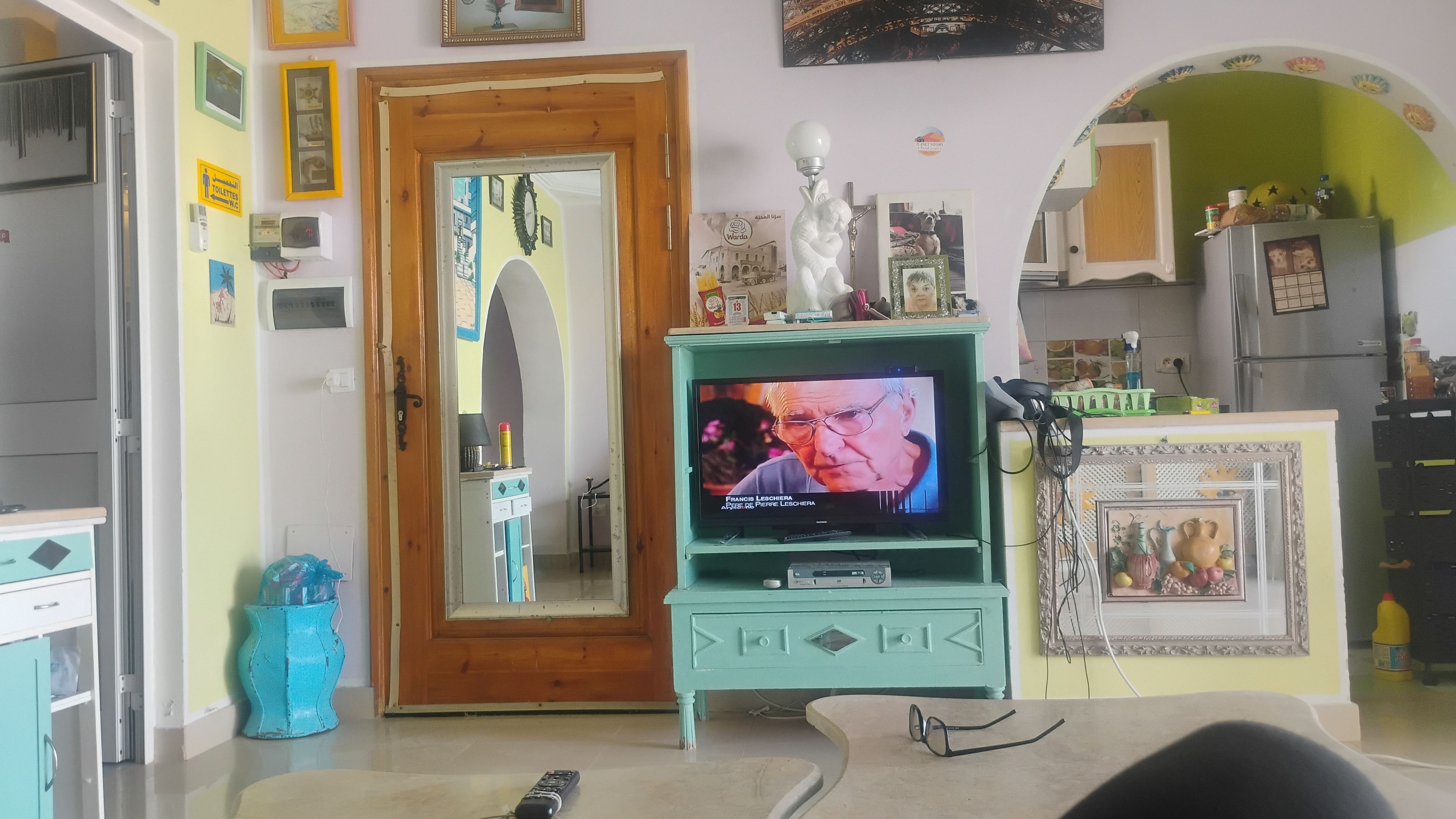 Sala de estar com televisão, uma porta e cozinha ao fundo.