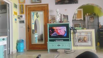 Sala de estar com televisão, uma porta e cozinha ao fundo.