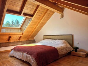 Quarto com teto inclinado, uma janela grande, cama com manta vermelha e mesa de cabeceira com luminária.