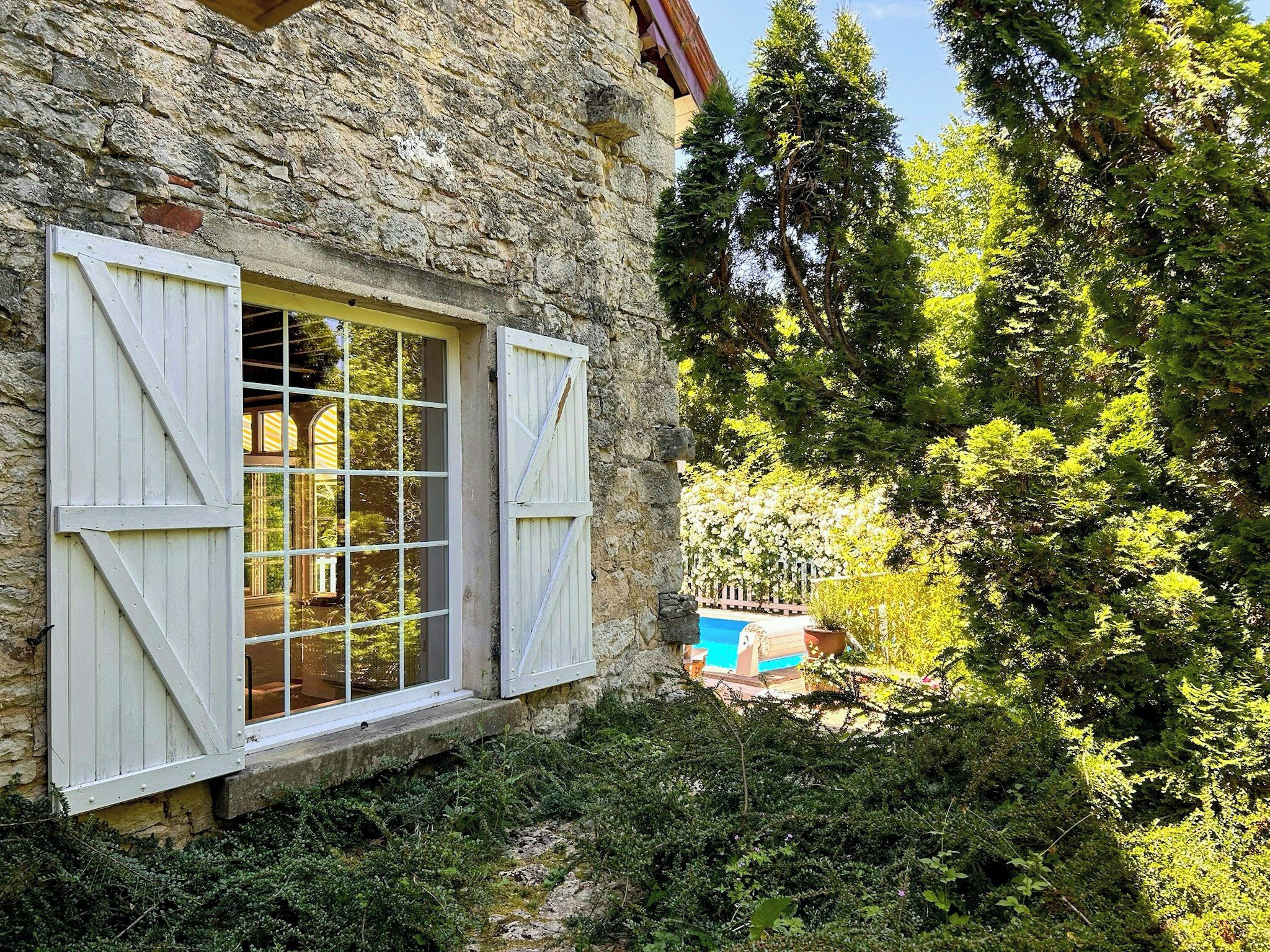 Un bâtiment en pierre doté d’une fenêtre et de volets, entouré de verdure et bordé d’une piscine.