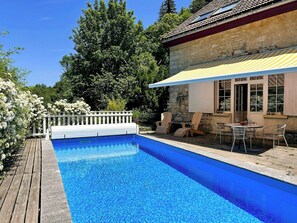 Piscina com deck de madeira, cerca branca e edifício de pedra com toldo amarelo.