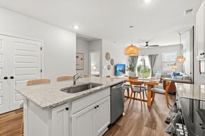 Una cocina moderna con un lavamanos integrado, un comedor con mesa y sillas de madera, y una sala de estar con sofá y televisor.