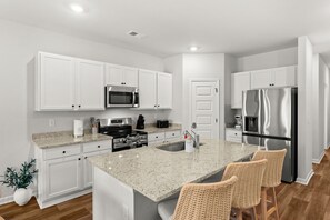 Cocina moderna con gabinetes blancos, electrodomésticos de acero inoxidable, encimera de granito y una zona de bar con sillas de mimbre.