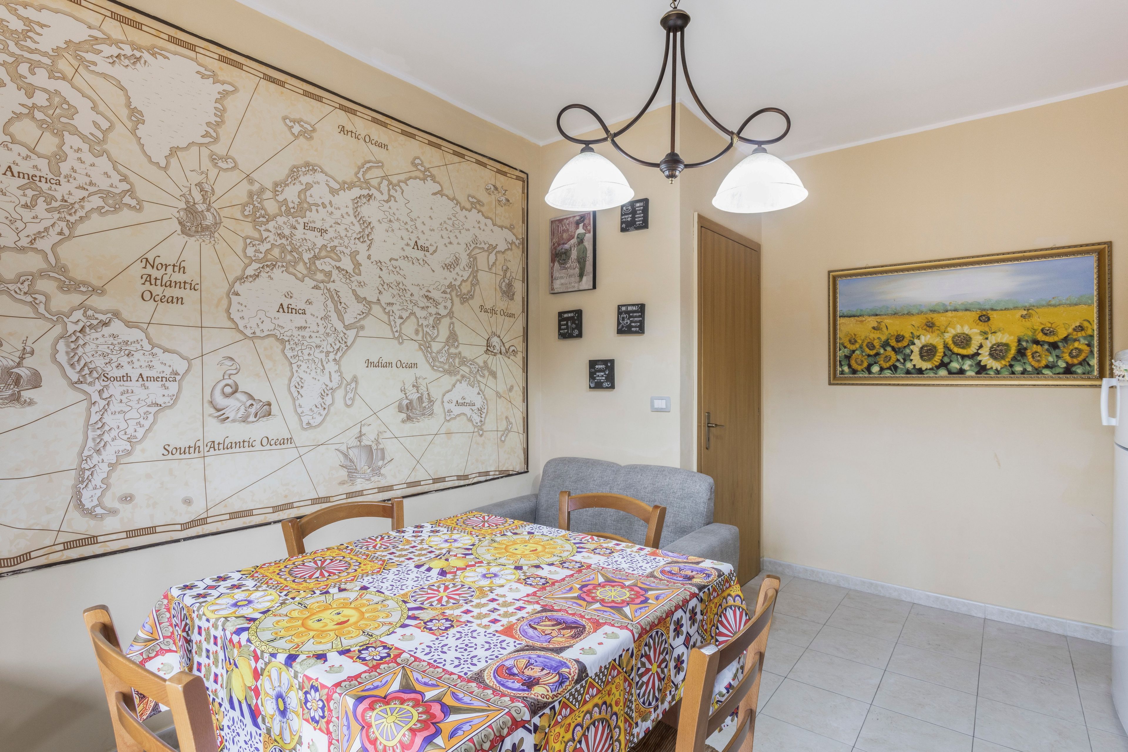 Een eethoek met een grote wereldkaart, een tafel met een kleurrijke tafelkleed en een zonnebloemen schilderij aan de muur.