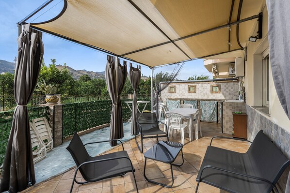 An outdoor patio with a table and chairs, a pergola with curtains, and a view of the surrounding landscape.