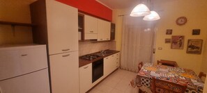 A compact kitchen with a refrigerator, stove, and cabinets, a dining table with a patterned tablecloth, and a wall clock.