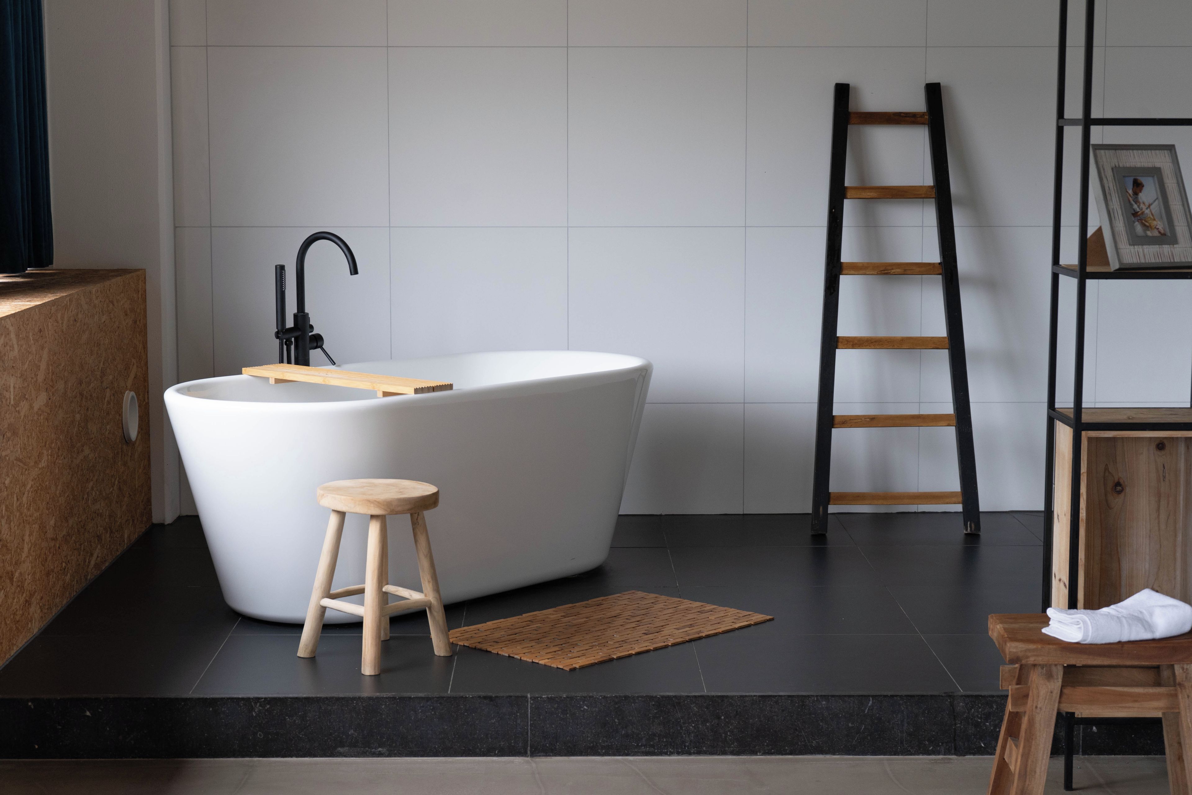 Een moderne badkamer met een vrijstaand bad, een houten kruk, een zwart betegelde vloer en een houten trap.