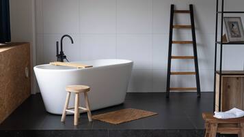 Een moderne badkamer met een vrijstaand bad, een houten kruk, een zwart betegelde vloer en een houten trap.