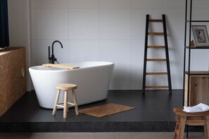 Ein modernes Badezimmer mit einer freistehenden Badewanne, einem hölzernen Hocker, einem schwarzen Fliesenboden und einer Holzleiter.