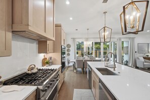 A modern kitchen with stainless steel appliances, a dining area with a white table and chairs, and a large window providing a view of the outdoors. - Golf Cart, Large Pool, Hot Tub, Walk 2 Beach, Bikes (Santa Rosa Beach)