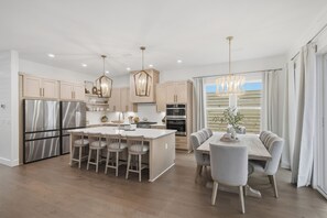 A modern kitchen with a central island, stainless steel appliances, and a dining area with a table and chairs. - Golf Cart, Large Pool, Hot Tub, Walk 2 Beach, Bikes (Santa Rosa Beach)