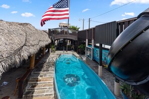 A swimming pool with a thatched roof structure on one side, a basketball hoop, and an American flag. - Crab Shack Boat Resort 5-Unit Tiki Resort On Water (Bay City)