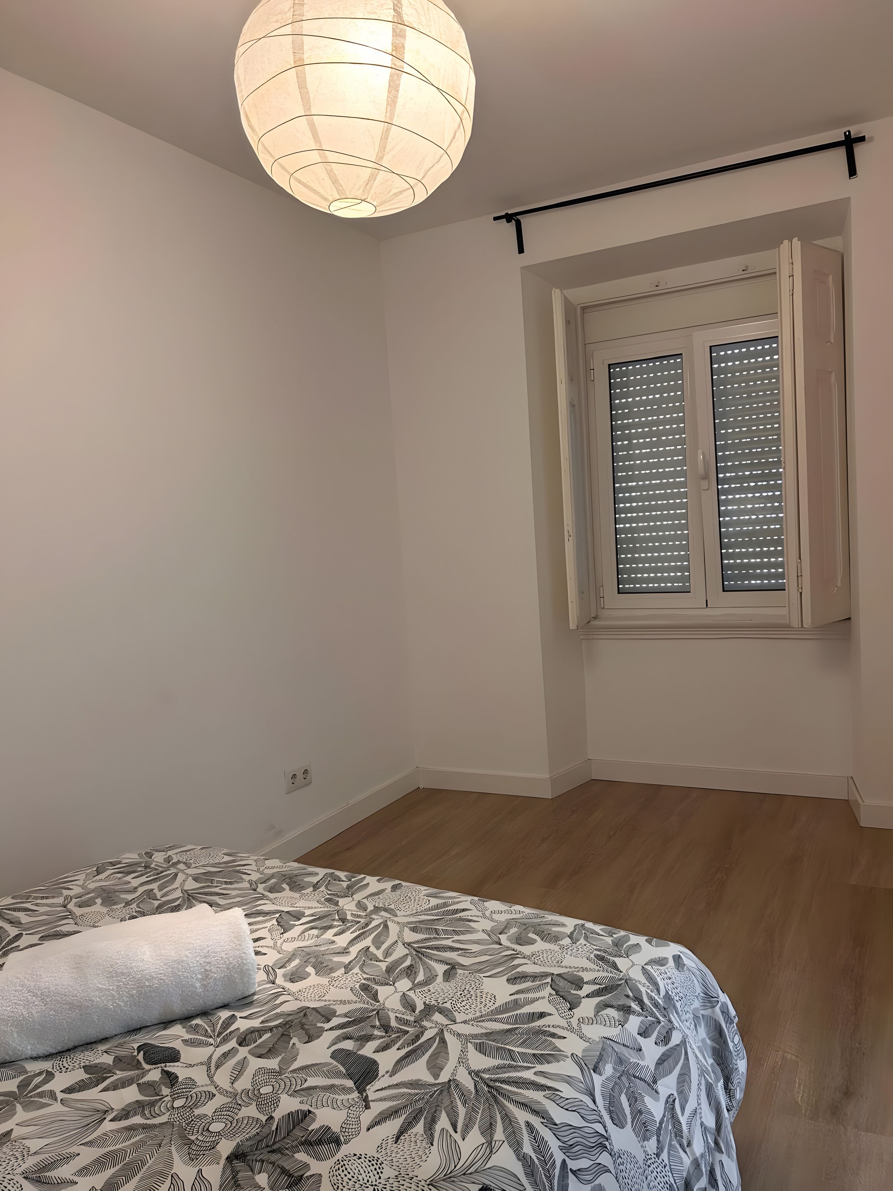 Ett sovrum med en säng, ett fönster med persienner, en hängande lampa och en dörr.