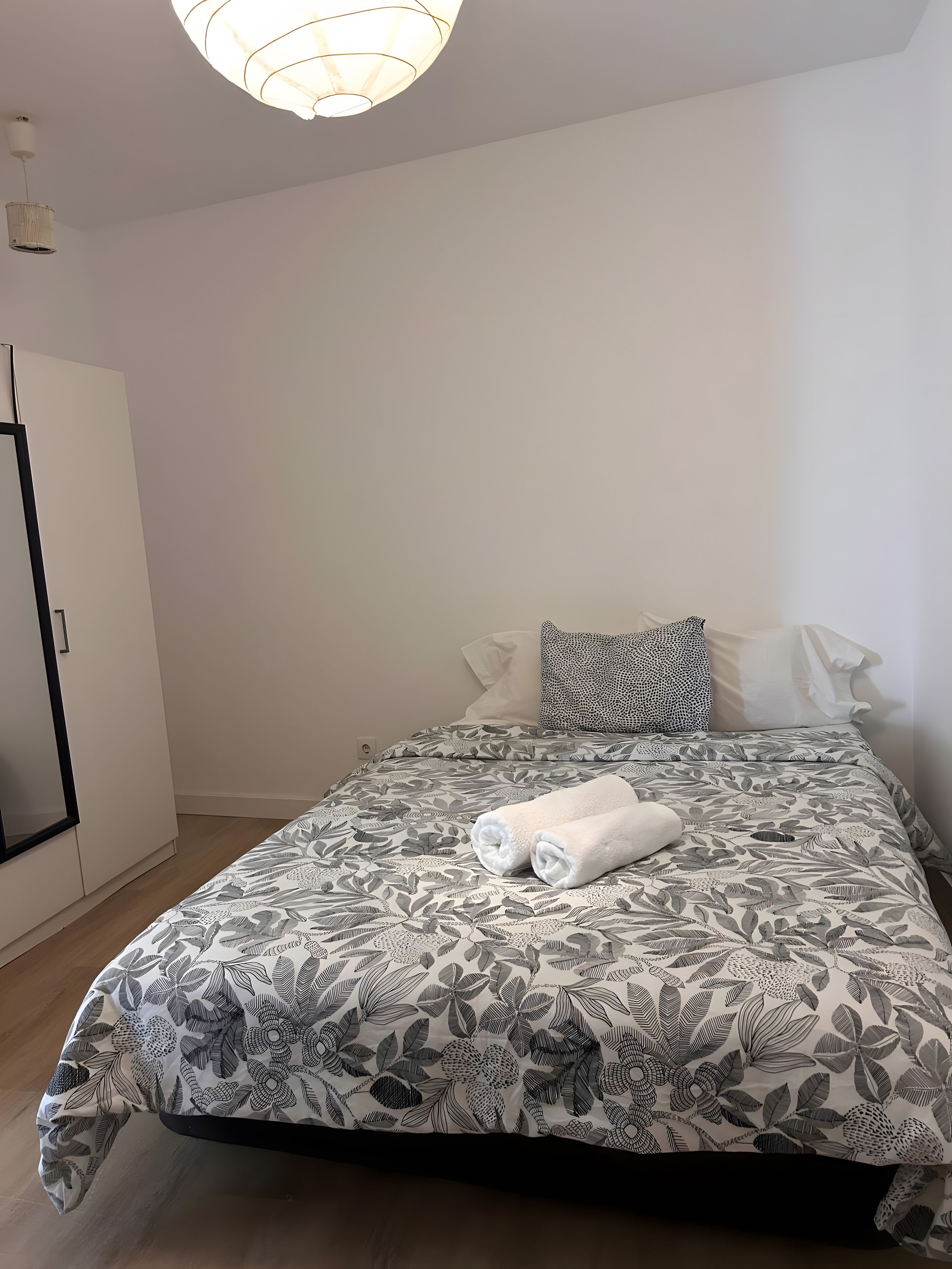 A neatly made bed with a floral-patterned bedspread, a rolled white towel, and a pillow with a geometric pattern.