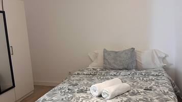 A neatly made bed with a floral-patterned bedspread, a rolled white towel, and a pillow with a geometric pattern.