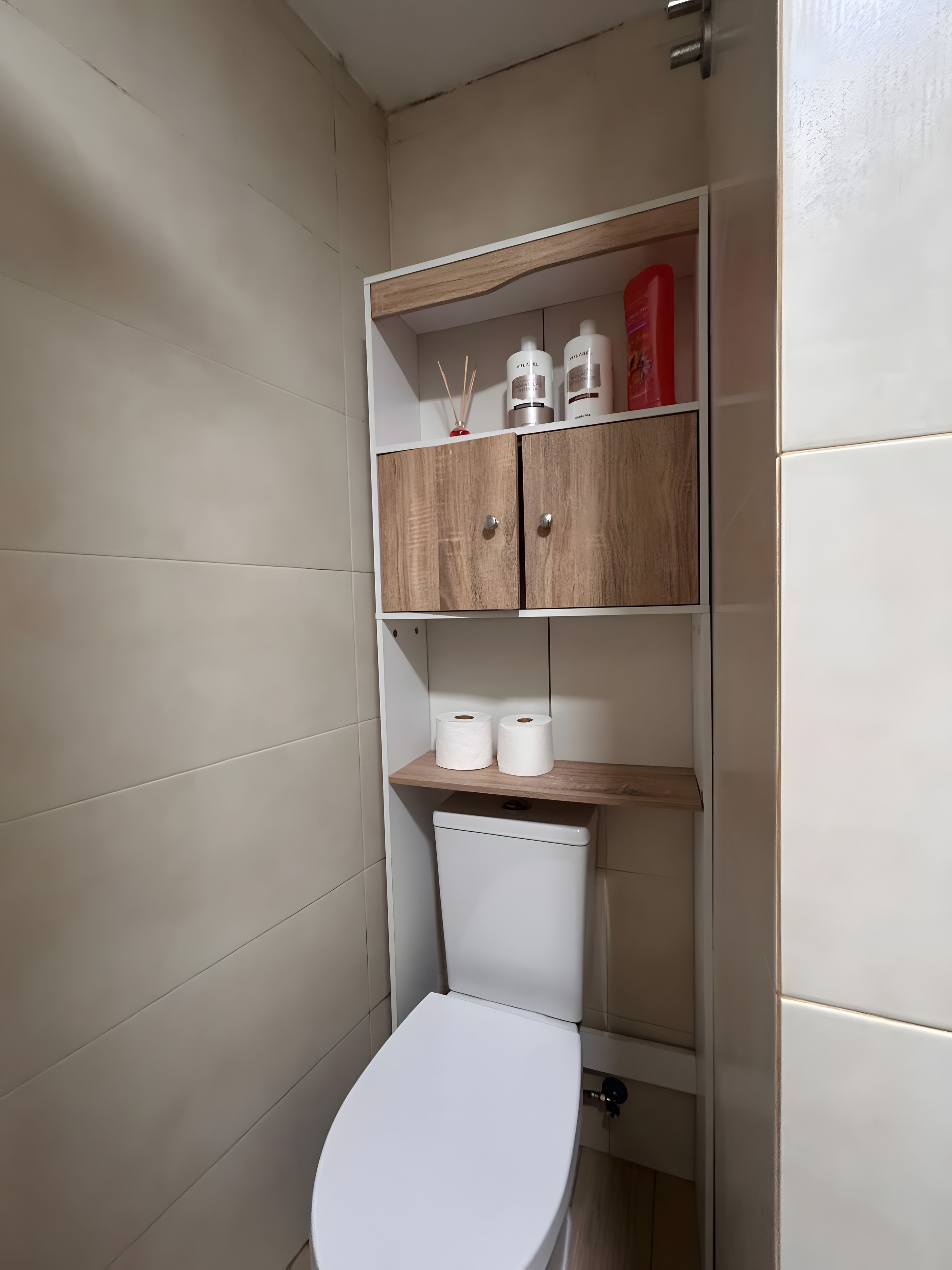 A bathroom with a white toilet, a wooden shelf with toiletries, and a white tiled wall.