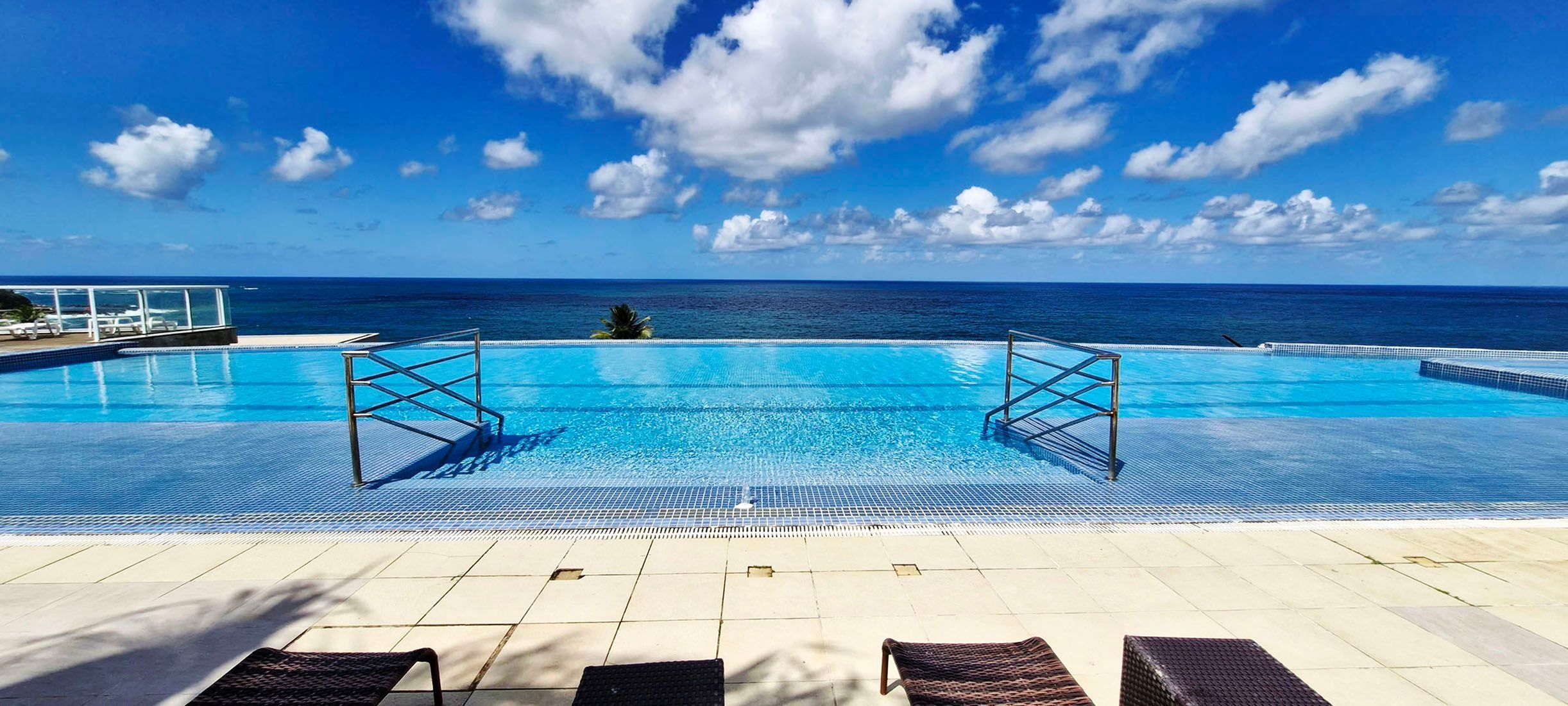 Céu azul claro com nuvens, piscina com guarda-corpo de metal e espreguiçadeiras.