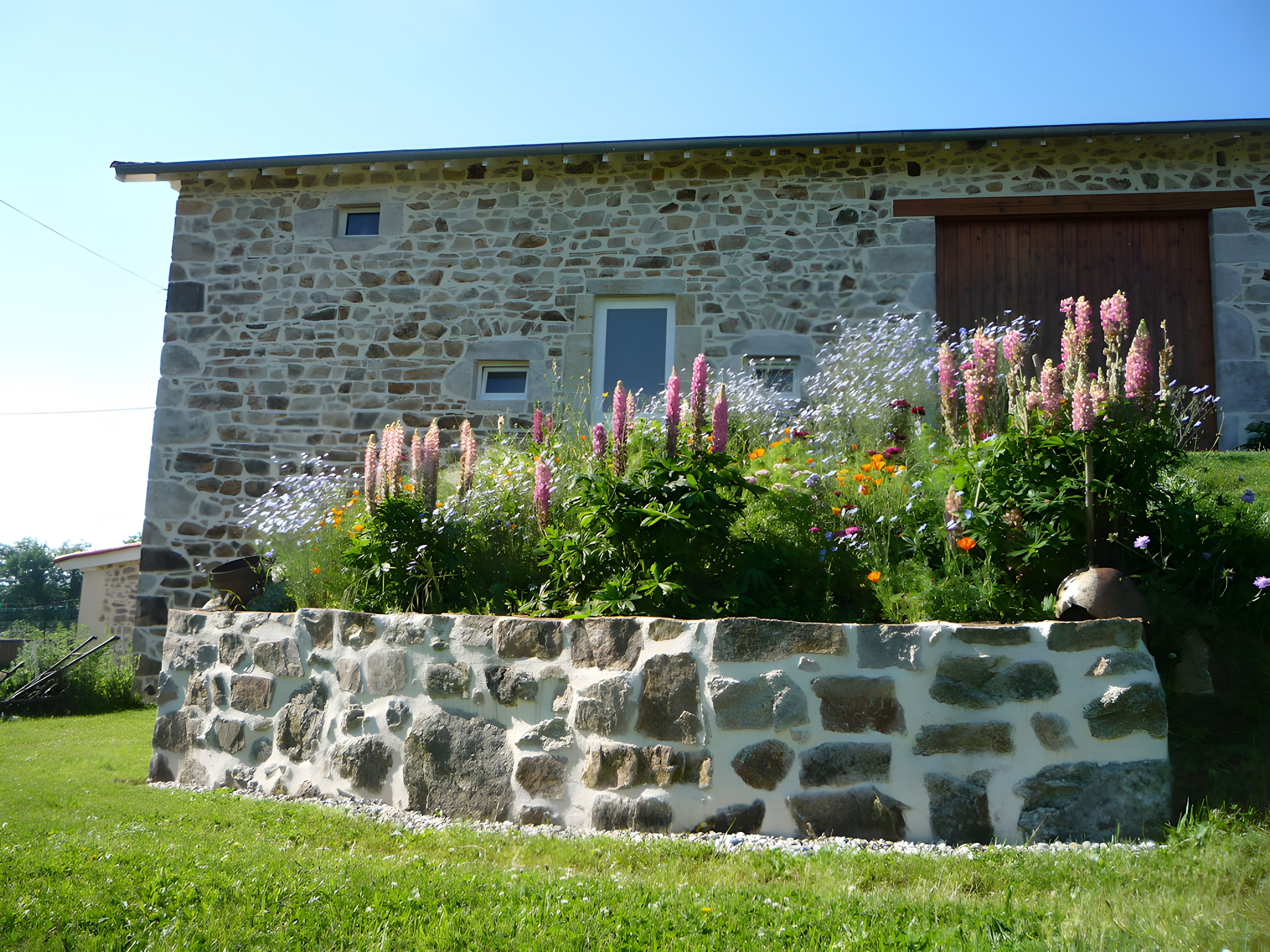 Un bâtiment en pierre doté d’une porte et de fenêtres en bois, entouré d’un mur de pierre et agrémenté de diverses plantes à fleurs.