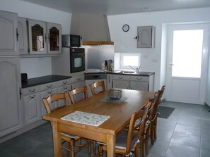 A dining area with a wooden table and chairs, a kitchen with white cabinets, a microwave, and a clock on the wall. - Holiday Home 'Les Hirondelles' (Saint-Georges-Lagricol)