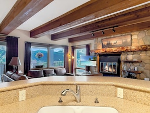A cozy living room with a stone fireplace, a large window with a scenic view, and a kitchen area with a granite countertop and sink. - New Listing - The Plaza 235, Walk to Crested Butte Lifts (Crested Butte)