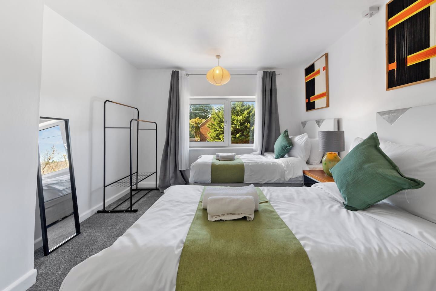 A hotel room with two beds, a window with a view of greenery, a hanging light fixture, and a mirror on the wall.