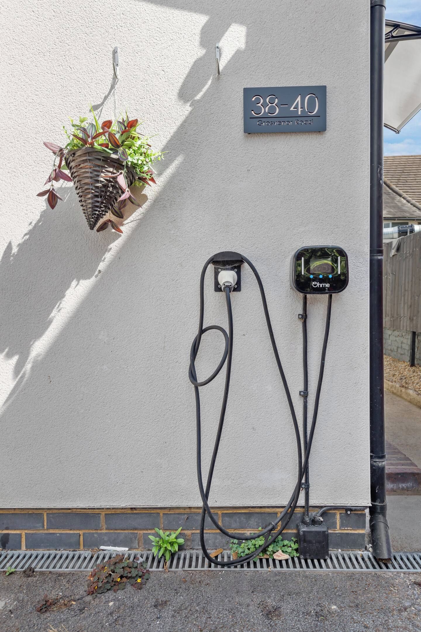 Estación de carga para vehículos eléctricos instalada en la pared, con un enchufe y cable visibles, una maceta colgante y un cartel con el número de la casa.