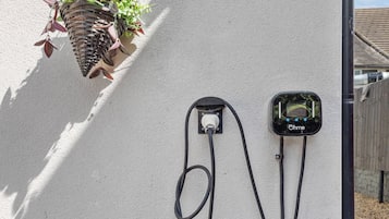 A wall-mounted electric vehicle charging station with a visible plug and cable, a hanging plant basket, and a house number sign.