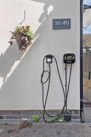 Estación de carga para vehículos eléctricos instalada en la pared, con un enchufe y cable visibles, una maceta colgante y un cartel con el número de la casa.