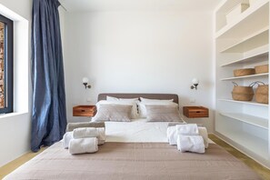 A bedroom with a bed, bedside tables, a bedside lamp, a window with a curtain, and a shelf with baskets. - Villa Marevento by Perle di Puglia (Carovigno)