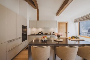 A modern kitchen with a central island, wooden beams, and a view of a cityscape through a window. - Paradisa Summit - Luxurious penthouse apartment with Mont-Blanc views (Chamonix-Mont-Blanc)