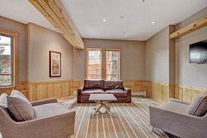 Un salon spacieux avec un canapé en cuir, deux fauteuils, une table basse, des poutres en bois, une grande fenêtre et un téléviseur fixé au mur.