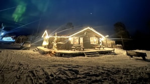 Uma casa com chão coberto de neve, um pinheiro de Natal iluminado e vista para o céu noturno com auroras boreais.