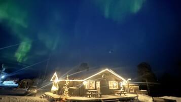 Une maison dont le sol est recouvert de neige, un sapin de Noël illuminé et une vue sur le ciel nocturne, illuminé par des aurores boréales.