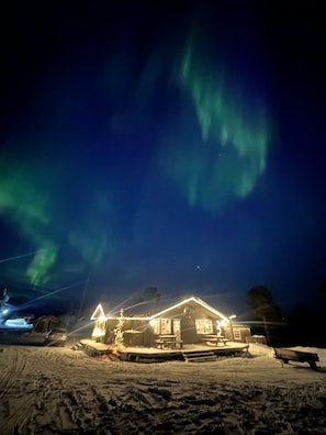 Una casa con el suelo cubierto de nieve, un árbol de Navidad iluminado y una vista del cielo nocturno con auroras boreales.
