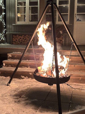 A fire pit with flames, surrounded by a wooden deck and a house in the background.